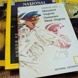 May include: A vintage National Geographic magazine cover featuring a close-up of a man in a colonial-era hat playing a flute. The cover text reads "CLOSE-UP USA" and lists the states of Maryland, Virginia, Delaware, and West Virginia. The magazine's logo is visible in the bottom right corner.