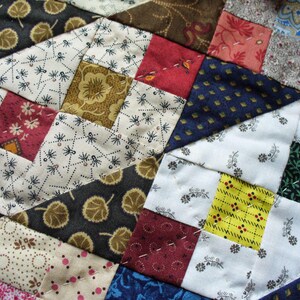 May include: Close-up of a patchwork quilt with a variety of patterned fabrics. The quilt features squares and triangles in shades of red, brown, blue, yellow, and white. The fabrics include floral, leaf, and geometric designs, stitched together with white thread.