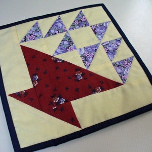 May include: A yellow and red patchwork quilt square with a navy blue border. The quilt square is made up of various triangles and squares in different patterns and colors. The center of the square features a large red arrow shape.