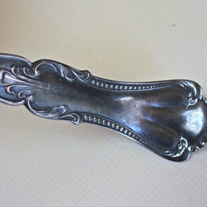 May include: Close-up of a silver-colored utensil handle with ornate scrollwork and a beaded border. The handle has a curved shape and a slightly tarnished appearance, suggesting age or use. The design is symmetrical and detailed.