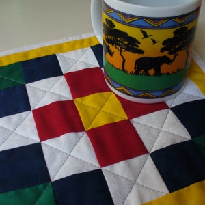 May include: A colorful patchwork quilt coaster with a yellow border. The coaster is made of squares of red, white, blue, green, and yellow fabric. A white mug with a colorful African animal design is sitting on the coaster.