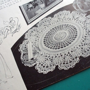 May include: A white crocheted doily with a delicate lace pattern. The doily is displayed on a black surface with a small, clear glass dish.