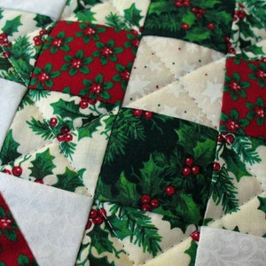 May include: Close-up of a quilted Christmas-themed fabric. The design features a patchwork pattern with red holly berries and green leaves on a white background. The quilting stitches create a diamond pattern.