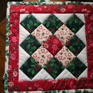 May include: A square Christmas-themed quilted potholder. The center features a diamond pattern of green holly, candy canes, and red poinsettias. A red floral border frames the design, with a white inner border. The potholder is designed for kitchen use.