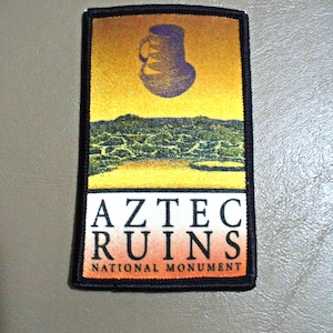 May include: A rectangular embroidered patch with a black border. The design features a purple pottery vessel above a green and yellow landscape. Below this, the text AZTEC RUINS is printed in black, followed by NATIONAL MONUMENT in smaller text.