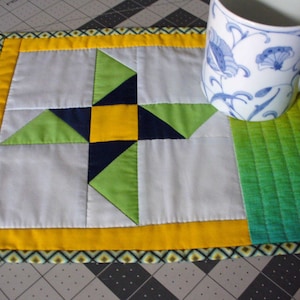 May include: A yellow, green, and blue quilted coaster with a star design. The coaster has a white border and a green and yellow border.