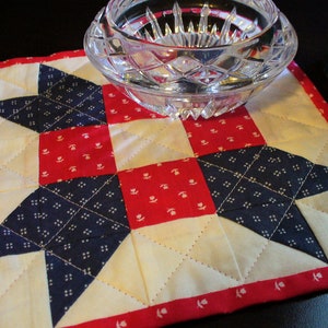 May include: A red, white, and blue quilted fabric coaster with a star pattern. The coaster is made of cotton fabric and is quilted with a white thread. The coaster is perfect for protecting surfaces from hot dishes or drinks.