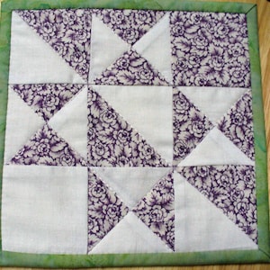 May include: A square quilted coaster with a green border and a pattern of white and purple triangles. The purple triangles have a floral print.