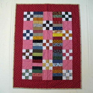 May include: A pink and red patchwork quilt with a variety of colorful fabric squares. The quilt is framed with a red border and has a white background.