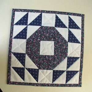 May include: A square quilt block with a central floral print surrounded by eight white triangles with navy blue borders. The quilt block is framed with a navy blue and white floral border.