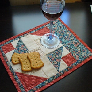 May include: A red, blue, and white quilted placemat with a glass of red wine and three crackers on top.