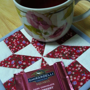 May include: A pink and white floral patterned mug filled with dark tea sits on a red and white patchwork quilt. A pink and white chocolate bar with the text "Ghirardelli Chocolate Squares Dark Chocolate Raspberry Luscious Filling" is on the quilt.