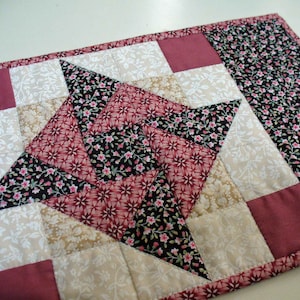 May include: A quilted table runner with a starburst design in shades of pink, brown, and white. The runner is made of fabric with floral patterns.