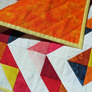 May include: A colorful quilt with a geometric pattern of triangles in shades of red, orange, yellow, blue, and white. The quilt is made with different fabrics and textures, creating a visually interesting and unique design.