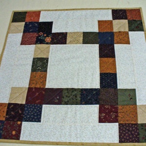 May include: A patchwork quilt with a white background and squares of various colors and patterns, including brown, green, red, and blue. The quilt is edged with a tan border.