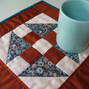 May include: A teal blue ceramic mug sits on a brown and white quilted coaster with a teal blue border. The coaster is made of fabric squares sewn together in a geometric pattern.