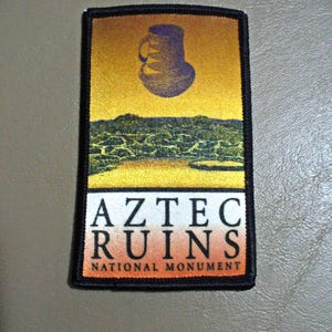 May include: Rectangular patch featuring a purple Aztec pot over a landscape with the text "AZTEC RUINS NATIONAL MONUMENT". The patch has a black border and a gradient background of yellow and orange.