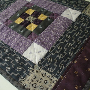 May include: A quilt made of various fabrics in shades of purple, gray, and white. The quilt is made up of squares and rectangles, and the fabric patterns include small floral prints, geometric patterns, and solid colors.