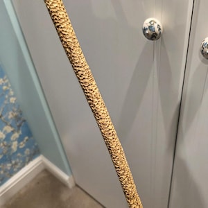 Op de afbeelding: Een getextureerde houten wandelstok met een koperkleurige metalen punt. Het oppervlak van de stok heeft een gebeeldhouwd, honingraatachtig patroon. De stok staat schuin voor een witte deur.
