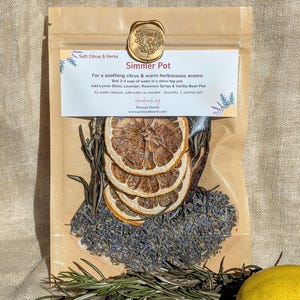 May include: A brown paper bag labelled "Simmer Pot" containing dried orange slices, lavender, and rosemary. The label says "Soft Citrus & Herbs" and describes the contents for a soothing aroma. A fresh lemon is visible.