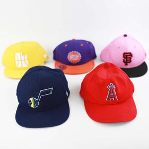 May include: Five baseball caps in various colors, including yellow, purple, pink, navy blue, and red. Each cap features a different team logo embroidered on the front. The caps have a flat brim and a structured crown.