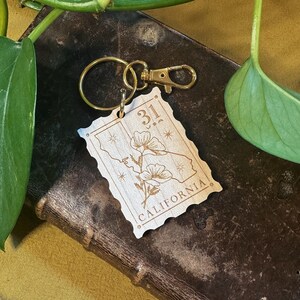 May include: Wooden keychain featuring a stylised outline of the state of California with a poppy flower design and the text "31" and "CALIFORNIA". The keychain has a gold metal ring and clasp.