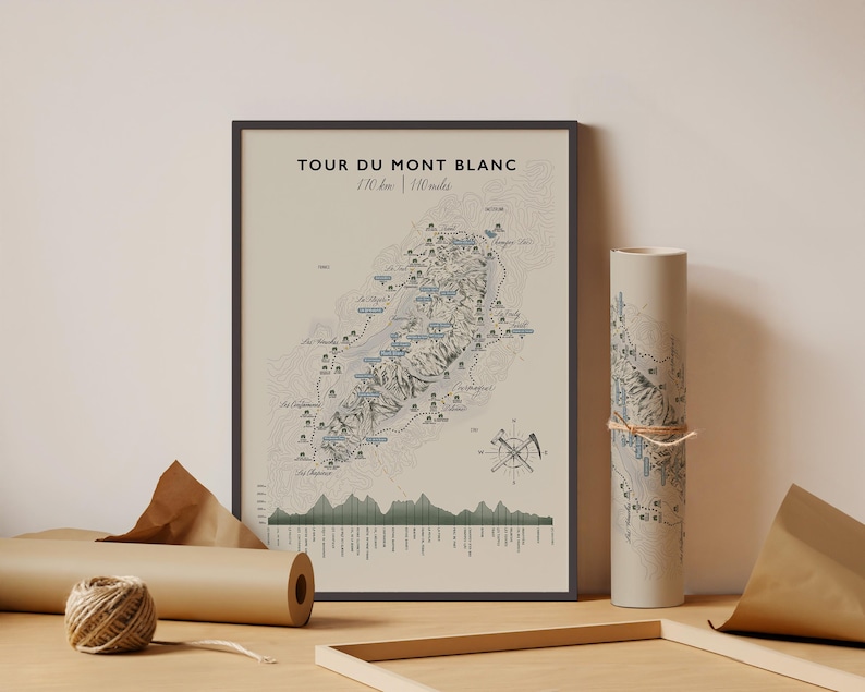 Op de afbeelding: Een ingelijste kaartprint met de titel TOUR DU MONT BLANC 110 km | 110 miles, met een topografische illustratie van de route, een windroos en een hoogteprofiel. Een opgerolde versie van dezelfde kaart is vastgebonden met een touwtje.