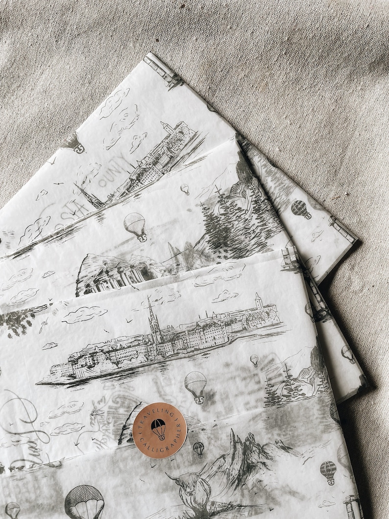 May include: A stack of white wrapping paper with a black and white city skyline and hot air balloon pattern. The top sheet of paper has a copper colored sticker with the text "Traveling Calligrapher" and a black hot air balloon graphic.
