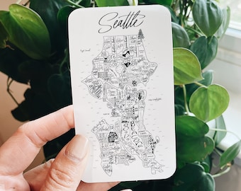 May include: A white card with a black and white illustration of a map of Seattle, Washington. The map is labeled with the names of different neighborhoods in Seattle. The card has the word "Seattle" written in black cursive at the top.