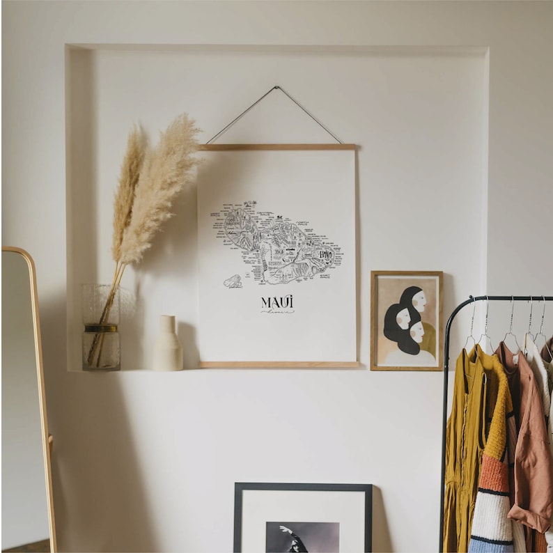 May include: A black and white line art print of a map of Maui, Hawaii. The print is framed in a light wood frame and hanging on a white wall. The print is titled "Maui" in black text.