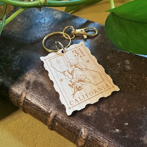 May include: Wooden keychain with a gold key ring. The keychain is shaped like a vintage postage stamp with the text "31" and "CALIFORNIA" on it. The stamp features a stylised image of a poppy flower.