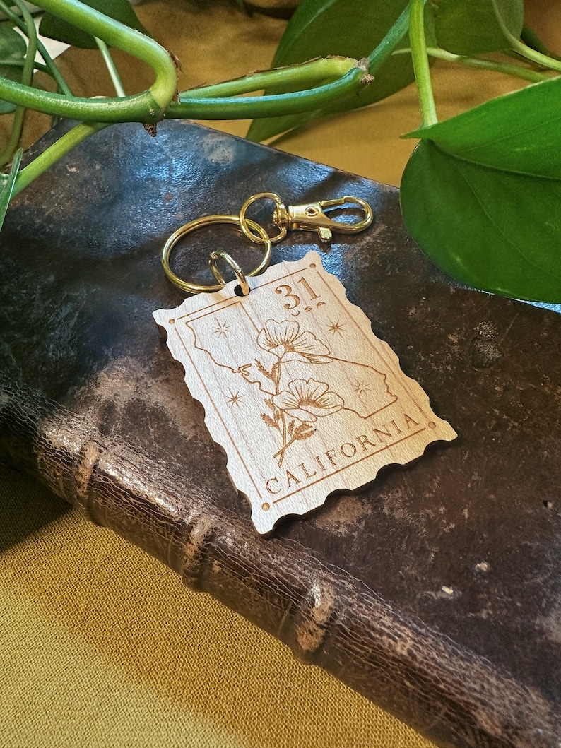May include: Wooden keychain with a gold key ring. The keychain is shaped like a vintage postage stamp with the text "31" and "CALIFORNIA" on it. The stamp features a stylised image of a poppy flower.