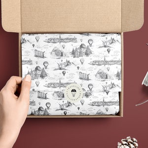May include: A white gift box with a brown cardboard lid is open, revealing tissue paper with a black and white pattern of hot air balloons and cityscapes. The tissue paper has a round sticker with the text "Traveling Calligrapher" in a circle.