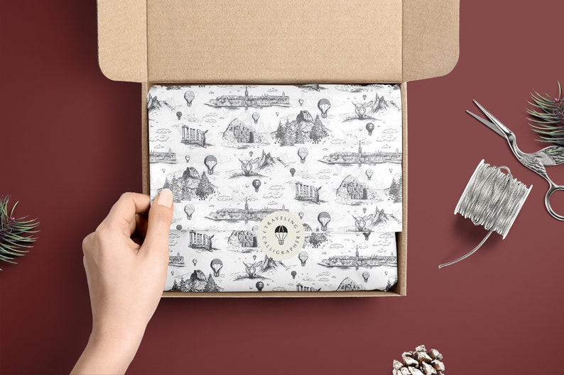 May include: A white gift box with a brown cardboard lid is open, revealing tissue paper with a black and white pattern of hot air balloons and cityscapes. The tissue paper has a round sticker with the text "Traveling Calligrapher" in a circle.