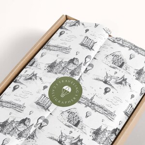 May include: White wrapping paper with a black and white pattern of hot air balloons, mountains, and buildings. A green circle with the text "Art Travelling Calligrapher" and a hot air balloon icon is in the center of the image.