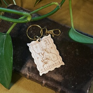 May include: A wooden keychain with a gold metal ring. The keychain features a stylised outline of the state of California with the number "31" and the word "CALIFORNIA" engraved on it. The keychain also features a small flower design.