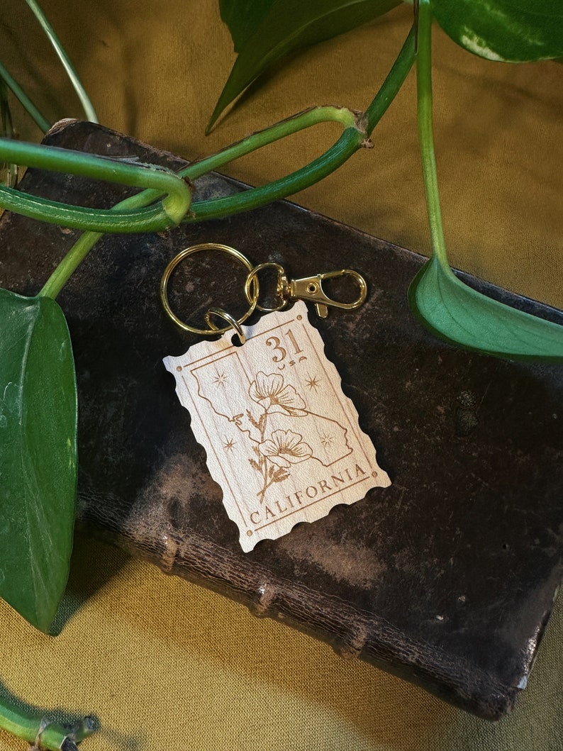 May include: A wooden keychain with a gold metal ring. The keychain features a stylised outline of the state of California with the number "31" and the word "CALIFORNIA" engraved on it. The keychain also features a small flower design.