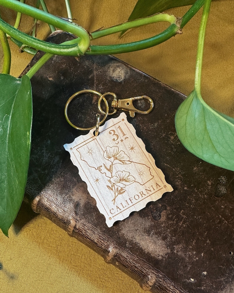May include: Wooden keychain featuring a stylised outline of the state of California with a poppy flower design and the text "31" and "CALIFORNIA". The keychain has a gold metal ring and clasp.