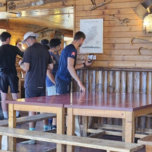 Op de afbeelding: Een houten tafel met een donkerrood oppervlak en een bijpassende houten bank in een kamer met houten wandpanelen. Een witte poster met de titel Tour du Mont Blanc hangt aan de muur en iemand gebruikt een smartphone om een label te scannen.