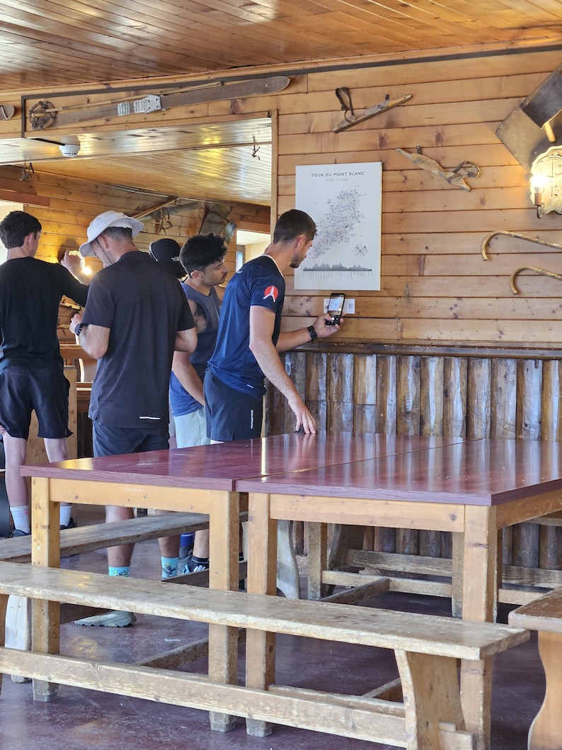 Op de afbeelding: Een houten tafel met een donkerrood oppervlak en een bijpassende houten bank in een kamer met houten wandpanelen. Een witte poster met de titel Tour du Mont Blanc hangt aan de muur en iemand gebruikt een smartphone om een label te scannen.