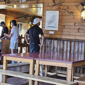Op de afbeelding: Een houten binnenruimte met twee grote rechthoekige tafels met roodbruine bladen en bijpassende houten banken. Een witte poster met de titel TOUR DU MONT BLANC is bevestigd aan de horizontale houten wand en toont een kaart en tekst.