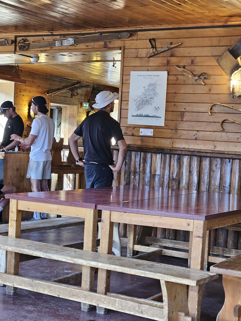 Op de afbeelding: Een houten binnenruimte met twee grote rechthoekige tafels met roodbruine bladen en bijpassende houten banken. Een witte poster met de titel TOUR DU MONT BLANC is bevestigd aan de horizontale houten wand en toont een kaart en tekst.