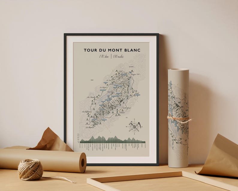 Op de afbeelding: Een ingelijste print getiteld TOUR DU MONT BLANC 170 km | 110 miles met een gedetailleerde topografische kaart van het bergpad, een hoogteprofiel en een kompasroos met ijsbijlen. Daarnaast staat een bijpassende opgerolde papieren print met touw.
