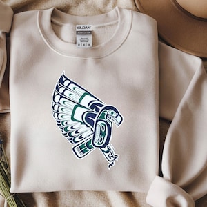 May include: A beige sweatshirt featuring a detailed graphic of a stylized eagle in shades of blue and green. The eagle design is centered on the front of the sweatshirt. The sweatshirt has a crew neck and long sleeves. The brand tag is visible at the top.