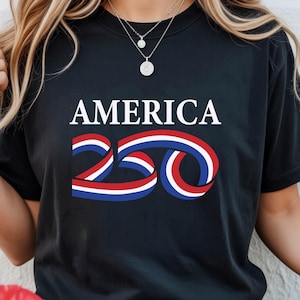 Puede incluir: Camiseta negra con la palabra "AMERICA" en blanco sobre un "250" estilizado en rojo, blanco y azul. El diseño tiene un tema patriótico.