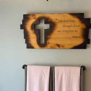 Puede incluir: Arte de pared de madera con un recorte de cruz y el texto "Todo lo puedo en Cristo que me fortalece." La madera tiene una veta natural con un borde oscuro y quemado. La obra de arte está montada en una pared gris claro.