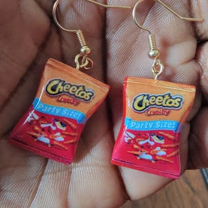 May include: A pair of dangle earrings featuring miniature replicas of Cheetos Crunchy Party Size snack bags. The charms are orange and red with the Cheetos logo and text printed on the front. They hang from gold-toned metal earring hooks.