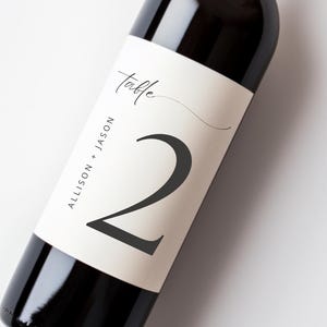 May include: A dark glass wine bottle featuring a cream-colored rectangular label. The label displays the word Table in script, the number 2 in a large dark font, and the text ALLISON + JASON printed vertically along the left side of the label.