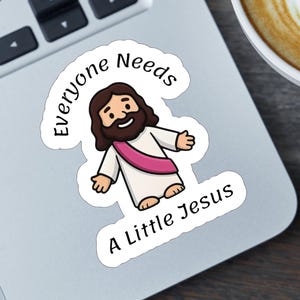 May include: A die-cut sticker featuring a cartoon illustration of a smiling figure with brown hair and a beard, wearing a white robe with a pink sash. The text "Everyone Needs A Little Jesus" is printed in black, curved around the central figure.