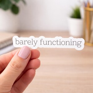 May include: A white sticker with the phrase "barely functioning" in a black, sans-serif font. The sticker is held by a hand with painted nails. The background includes a desk, a plant, and office supplies.
